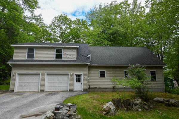 Two-story beige house with two white garage doors, back driveway, and surrounding green trees under a partly cloudy sky.