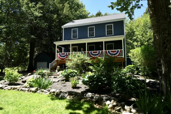 Blue two-story house with a wraparound porch decorated with red, white, and blue bunting in a lush garden. and trees surrounding the property.