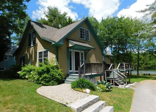 Yellow cottage with teal trim and a wooden deck by a lake on a sunny day, surrounded by trees.